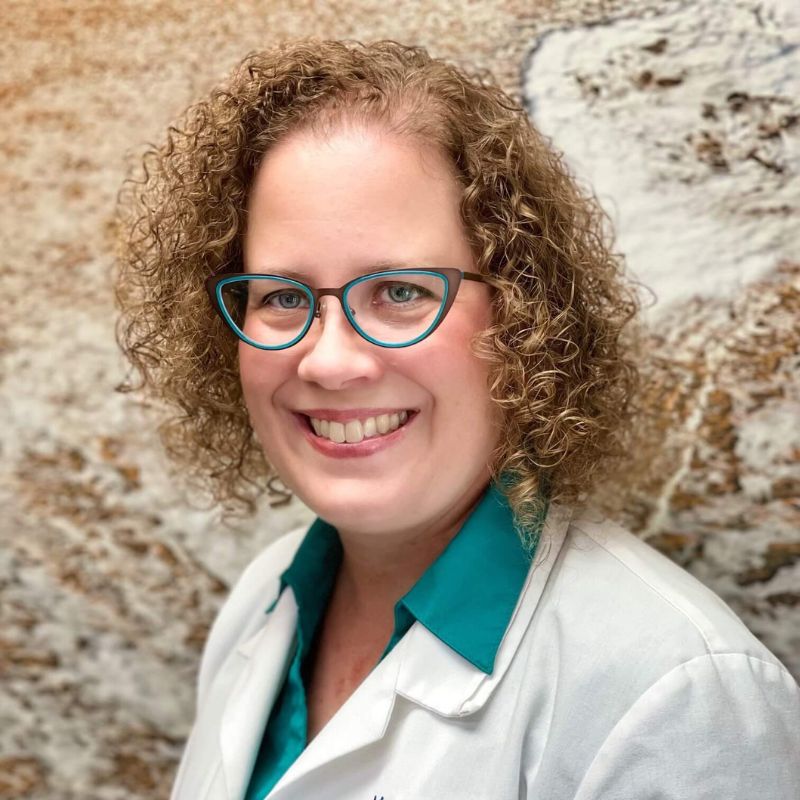 Dr. Marni Novick, audiologist, smiling in teal glasses and a white lab coat over a teal shirt